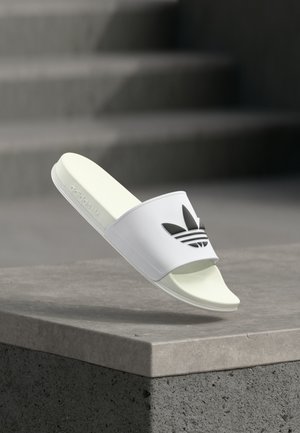 Balta Adidas slīduve ar melnu trefoil logotipu, novietota gaisā virs betona apmales, fonā redzamas izplūdušas betona kāpnes.