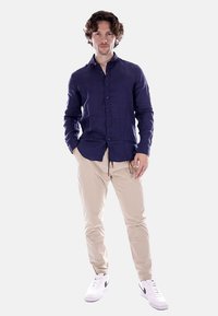 Maglietta button-up in lino blu navy, chinos beige e sneaker bianche con dettagli neri, in piedi su uno sfondo neutro.