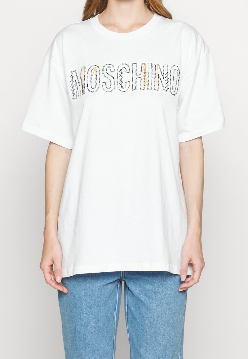Kobieta w białej, oversize'owej koszulce z naszytym logo "MOSCHINO" na wysokości klatki piersiowej oraz jasnoniebieskich dżinsach, stojąca na białym tle.