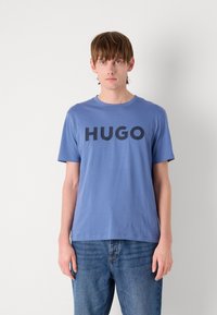 Mėlyna trumpomis rankovėmis medvilninė marškinėlė, su dideliu juodu "HUGO" logotipu priekyje ir apvaliu iškirpimu. Dėvimos su mėlynomis džinsais.