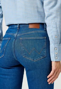 Vaqueros de mezclilla azul con un diseño ajustado, que presentan un parche de cuero de Wrangler y costuras en zigzag en los bolsillos traseros. Camisa de cuadros de manga larga.