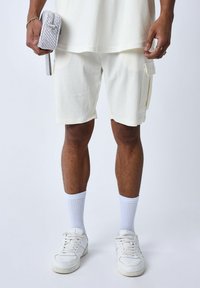 Pantalones cortos blancos con textura, bolsillos y acentos amarillos, combinados con calcetas blancas y zapatillas blancas. Una bolsa pequeña con estampado se sostiene en la mano.