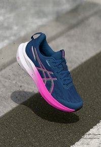 Μπλε και ροζ αθλητικό παπούτσι Asics σε θέση πτήσης πάνω από επιφάνεια ασφάλτου με λευκή γραμμή του δρόμου στο παρασκήνιο.