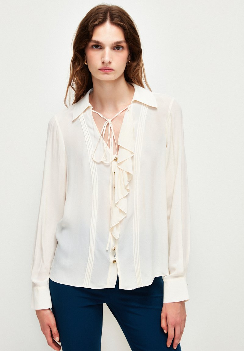 Blusa de seda blanca con un frente con volantes, mangas largas y un detalle de lazo en el cuello. Presenta costuras verticales y un corte holgado. Textura suave.