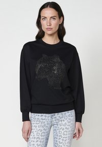 Felpa nera con un design di testa di leone realizzato in borchie d'argento. Maniche lunghe, polsini a costine e scollo classico. Abbinata a jeans con stampa leopardata chiara.