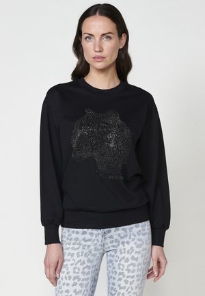 Felpa nera con un design di testa di leone realizzato in borchie d'argento. Maniche lunghe, polsini a costine e scollo classico. Abbinata a jeans con stampa leopardata chiara.