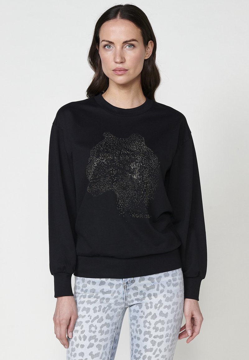 Felpa nera con un design di testa di leone realizzato in borchie d'argento. Maniche lunghe, polsini a costine e scollo classico. Abbinata a jeans con stampa leopardata chiara.