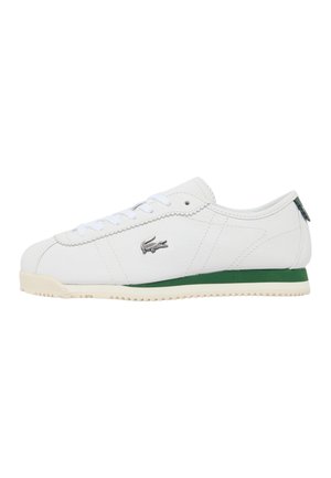 Zapatilla deportiva blanca de corte bajo con una franja verde en la suela, cordones blancos y un logo de cocodrilo plateado en el lateral.