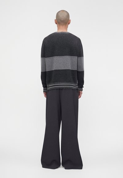 Personne à la tête rasée portant un pull rayé noir et gris et un pantalon noir à larges jambes, debout de dos sur un fond uni.