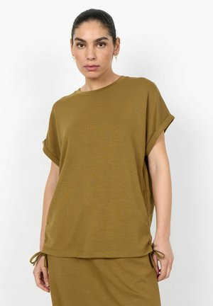 Femme portant un haut ample à manches courtes vert olive avec manches retroussées et jupe assortie, posant devant un fond blanc.