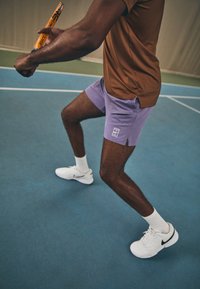 Lilla idrettsshorts med sidelommer, brun tettsittende skjorte og hvite joggesko, plassert på en blå idrettsbane. Tennisracket holdt i hånden.