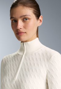 Lys cremefarvet pullover med høj krave, der har et struktureret diamantmønster og halv lynlås, lavet af blødt strikket materiale.