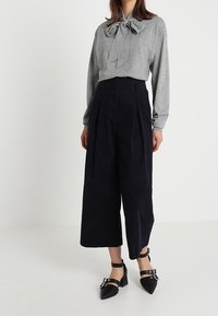 Un haut gris à manches longues avec un détail nœud papillon, associé à un pantalon large taille haute bleu marine et des escarpins noirs à lanières.