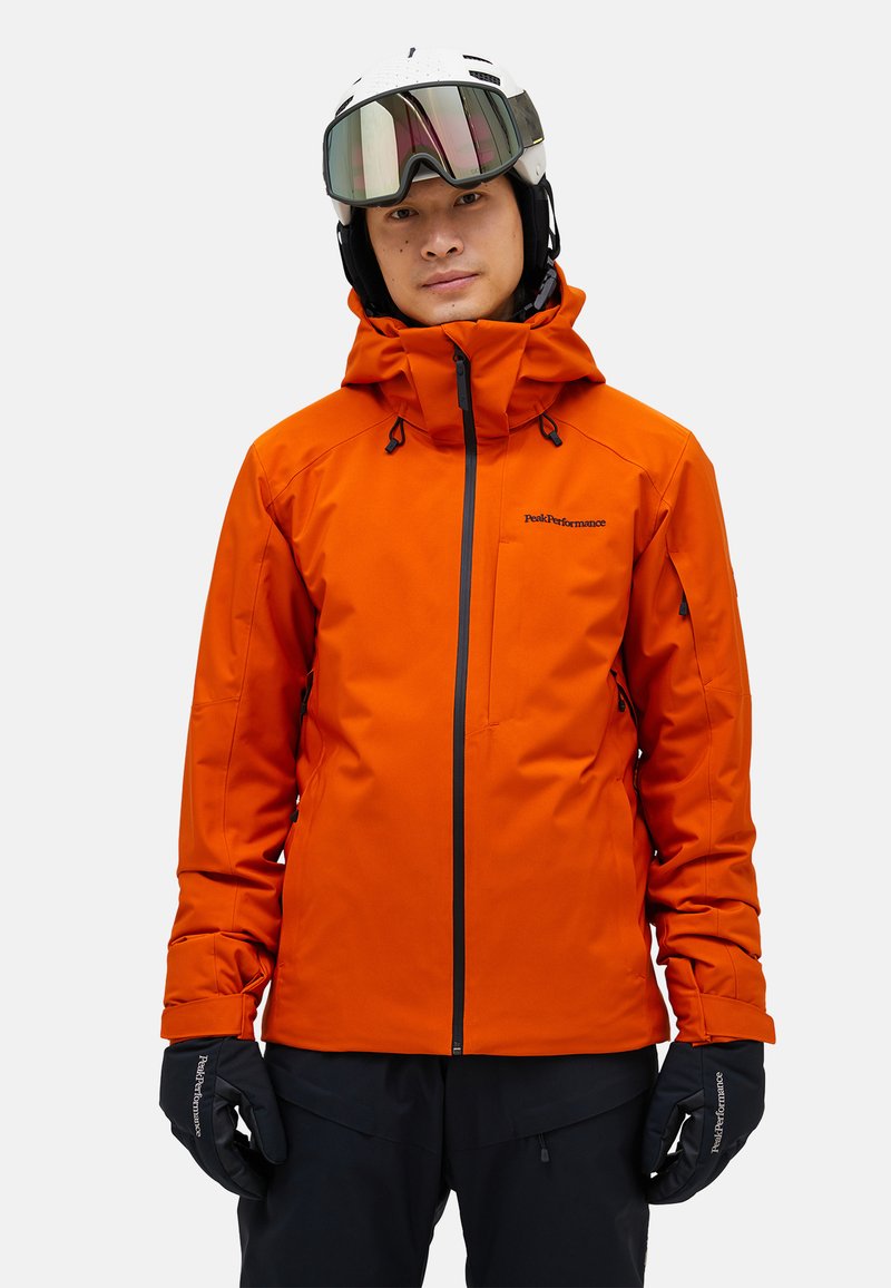 Lys orange skijakke med tætsiddende design, vandtæt materiale, justerbar hætte, sorte lynlåse og manchetter. Båret med handsker.
