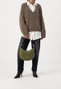 Person som bär en brun V-ringad tröja över en vit skjorta, mörka byxor, svarta klackboots, och håller en olivgrön handväska i handtaget.