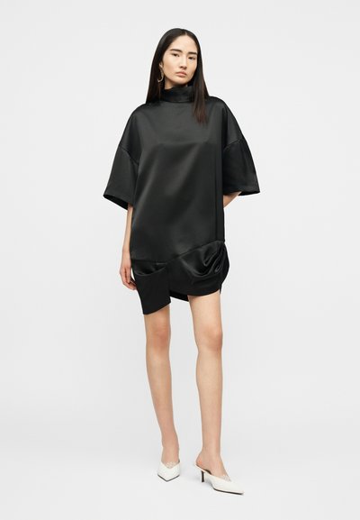 Γυναίκα με αέρινο μαύρο σατέν μίνι φόρεμα με ψηλό λαιμό και κοντά μανίκια, συνδυασμένο με λευκές μυτερές γόβες mule.