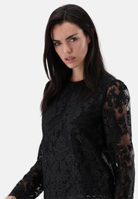 Schwarzes, florales Spitzentop mit transparenten langen Ärmeln und filigraner Stickerei. Der Stoff ist glatt mit strukturierten Mustern und einem runden Ausschnitt.