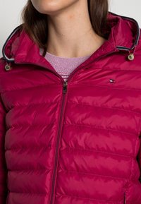 Fuksia toppatakki vetoketjulla edessä, jossa huppu, kontrastiväriset raidalliset reunat ja pieni logo rinnassa. Sileä tekstuuri.