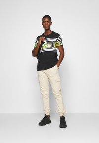 T-shirt noir avec des manches rayées et un panneau graphique coloré, associé à un pantalon cargo beige et des baskets noires.