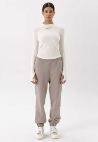 Wit ribbed langemouwen shirt met zwarte tekst, gecombineerd met lichtgrijze joggingbroek met elastische manchetten en een logo op het been, afgemaakt met witte sneakers.