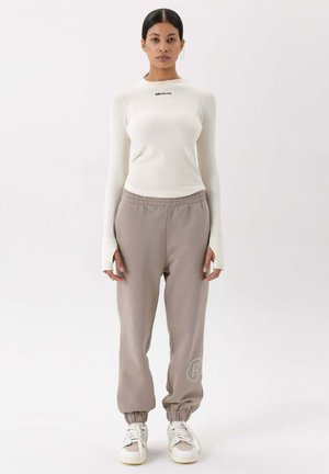 Wit ribbed langemouwen shirt met zwarte tekst, gecombineerd met lichtgrijze joggingbroek met elastische manchetten en een logo op het been, afgemaakt met witte sneakers.