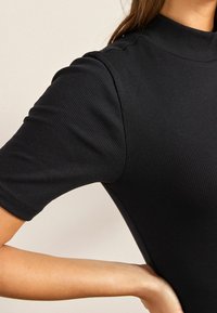 Haut noir à côtes avec un col montant et des manches courtes, présentant une coupe structurée et une texture lisse. Vue en gros plan de l'épaule et du bras.