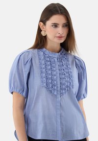 Hellblau gestreifte Bluse mit Puffärmeln, Rüschenverzierung in der Mitte und hohem Kragen. Hergestellt aus einem weichen, leichten Stoff.