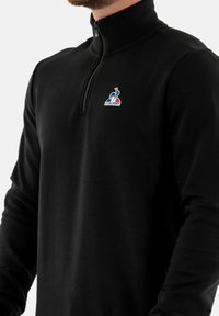 Zwarte zip-up sweatshirt met lange mouwen. Glad van structuur. Bevat een klein gekleurd logo op de linkerborst. Minimalistisch ontwerp.