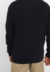 Marinblå ribbstickad tröja med långa ärm, rundad halsringning och en något längre bakre fåll. Texturen verkar mjuk och tjock.