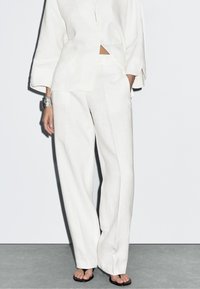 Vrouw in losse witte linnen broek en bijpassend knoopshirt met zilveren klokarmband en zwarte teenslippers.