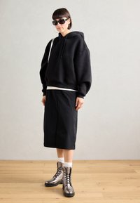 Un sweat à capuche noir surdimensionné avec un ourlet arrondi, associé à une jupe noire longue jusqu'aux genoux et des bottines à lacets métalliques. Accessoirisé avec des lunettes de soleil.