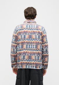 Άτομο που φοράει πολύχρωμο φλις μπουφάν με γεωμετρικά σχέδια, στέκεται με την πλάτη προς την κάμερα μπροστά σε ένα απλό φόντο.