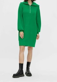 Robe tricotée verte avec un col zippé, des manches bouffantes longues et une coupe décontractée, associée à des bottines noires à semelle épaisse.