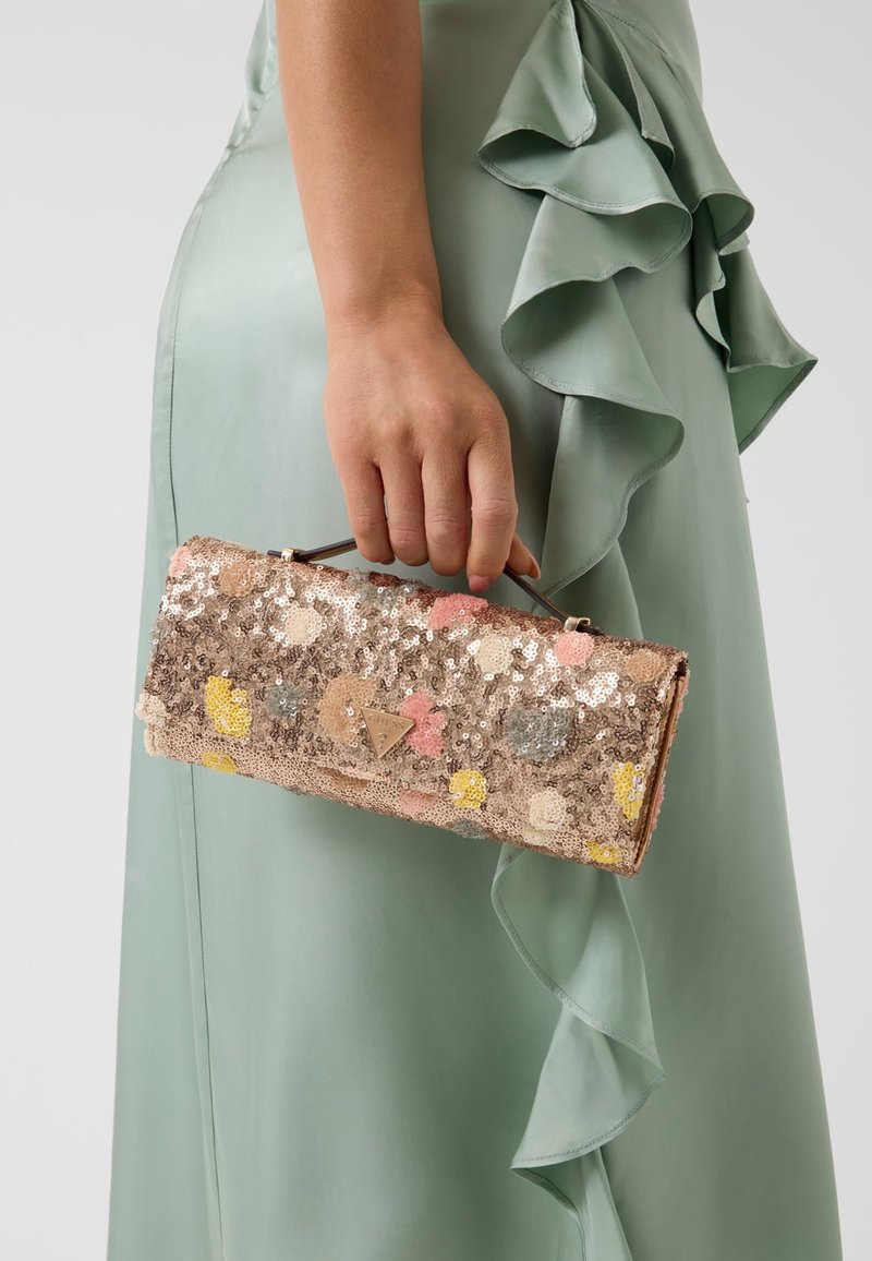 Femme tenant une pochette rectangulaire à sequins avec motifs floraux, portant une robe verte sauge à volants, vue de la taille vers le bas.
