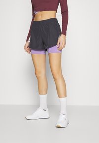 Musta urheiluhousut leveällä vyötärönauhalla laventelinväristen puristushousujen päällä, yhdistettyinä valkoisiin lenkkareihin ja nilkkasukkiin.