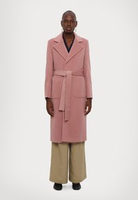 Lång rosa ullblandad kappa med slagkrage, bälte i midjan och två framfickor. Kombineras med vida beigefärgade byxor och svarta skor.