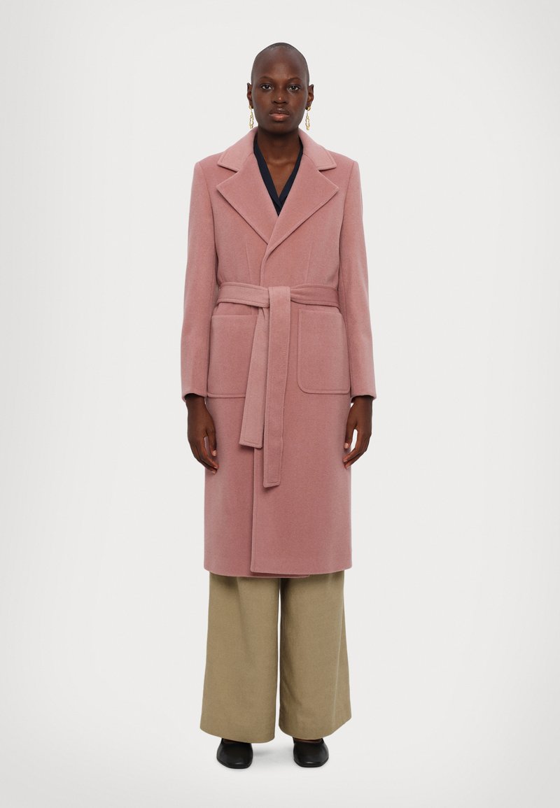 Lång rosa ullblandad kappa med slagkrage, bälte i midjan och två framfickor. Kombineras med vida beigefärgade byxor och svarta skor.