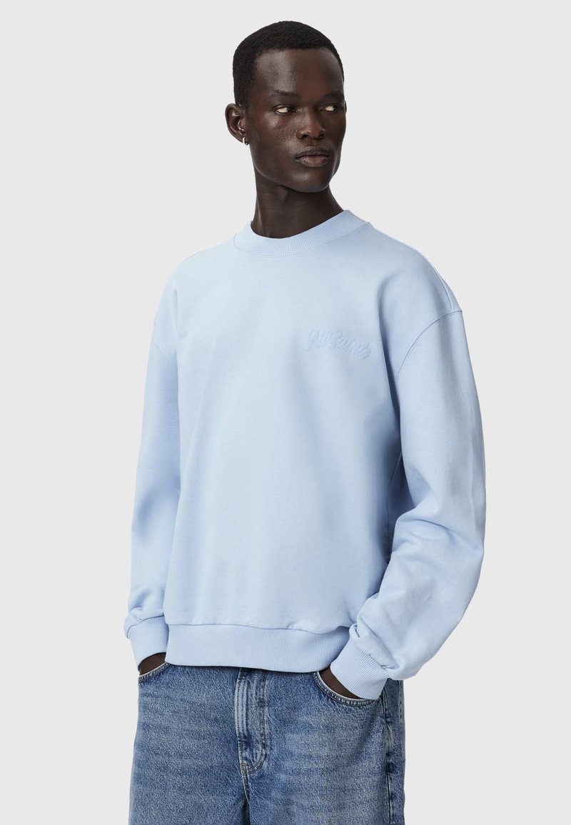 Homme à la peau foncée portant un sweat-shirt bleu clair et un jean bleu, mains dans les poches, regardant vers sa gauche sur un fond uni.