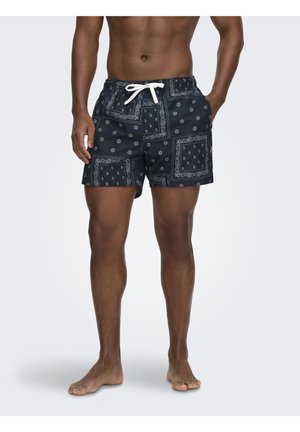 Hombre con pantalones cortos de baño negros con estampado blanco de bandana y cordón blanco, de pie descalzo con una mano en el bolsillo.