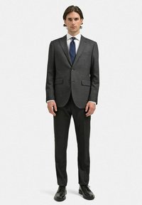 Mörkgrå skräddarsydd kostym med jacka med klyvkrage, enkel knappstängning och byxor med räta ben. Kombineras med en vit skjorta och marinblå slips.