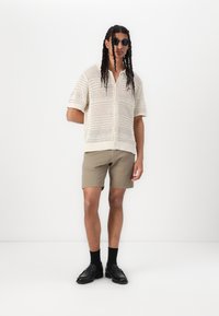 Lys beige, åbenstrikket skjorte med korte ærmer, sat sammen med khakifarvede shorts. Sorte sko og solbriller fuldender looket. Ensartet baggrund.
