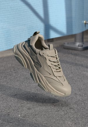 Beżowy masywny sneaker z grubą podeszwą i siateczkowymi detalami zawieszony nad szarym asfaltem na tle niebieskiej ściany.