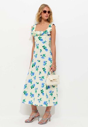 Mujer con vestido midi color crema con estampado floral azul, escote con volantes, gafas de sol, sujetando un pequeño bolso blanco y con tacones grises de tiras.