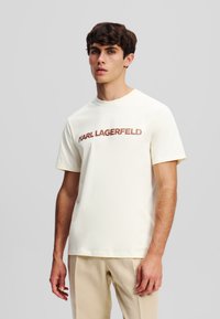 Crème-kleurige katoenen t-shirt met een ronde halslijn, korte mouwen en de tekst "KARL LAGERFELD" in bruine letters over de borst.