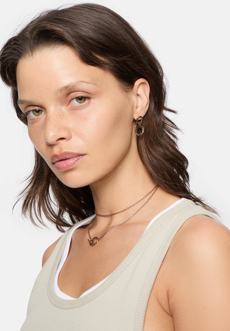 Goldfarbene Kettenhalskette mit einem Buchstabenanhänger, kombiniert mit einem beigen Tanktop mit weißem Saum. Die Person hat welliges braunes Haar.