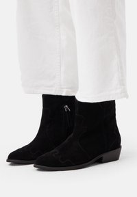 Stivali alla caviglia in suede nero con tacco basso, caratterizzati da motivi di cucitura curvi e una cerniera laterale. Abbinati a pantaloni bianchi a vita corta.