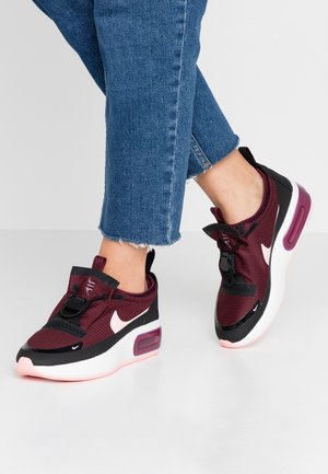 Persoon met donkerrode Nike-sneakers met witte swoosh en dikke witte zolen, gecombineerd met blauwe cropped jeans tegen een neutrale achtergrond.