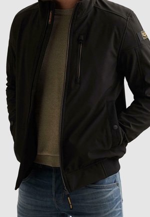 Homme portant une veste zippée noire par-dessus un pull en maille beige et un jean bleu, les mains dans les poches de la veste.