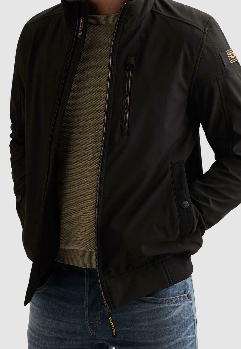Homme portant une veste zippée noire par-dessus un pull en maille beige et un jean bleu, les mains dans les poches de la veste.