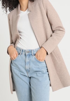Femme portant un manteau texturé beige clair par-dessus un haut blanc et un jean bleu clair taille haute, les mains dans les poches devant un fond uni.